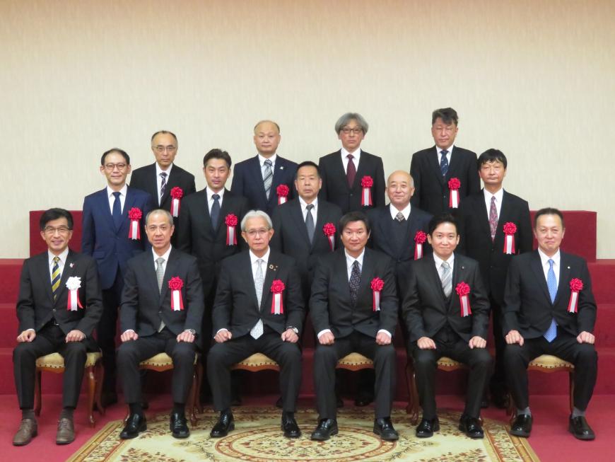 左胸に胸章リボンをつけた表彰された方が1列目に市長を含め6名が座り、2列目に5名、3列目に4名が立っている記念写真