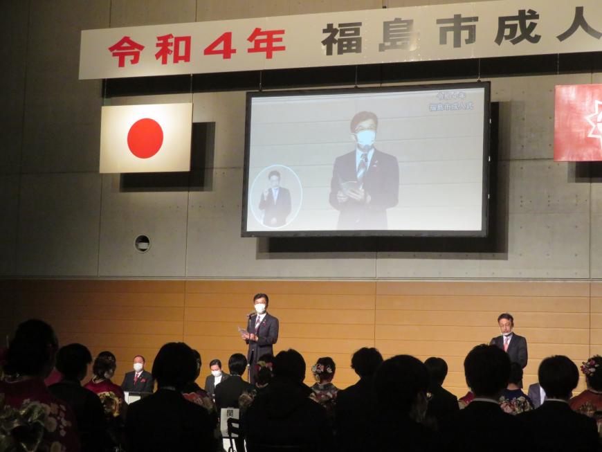 成人式の横断幕がある会場で、座っている参加者の前で、市長が立って話をしている様子がモニターに映し出されている写真