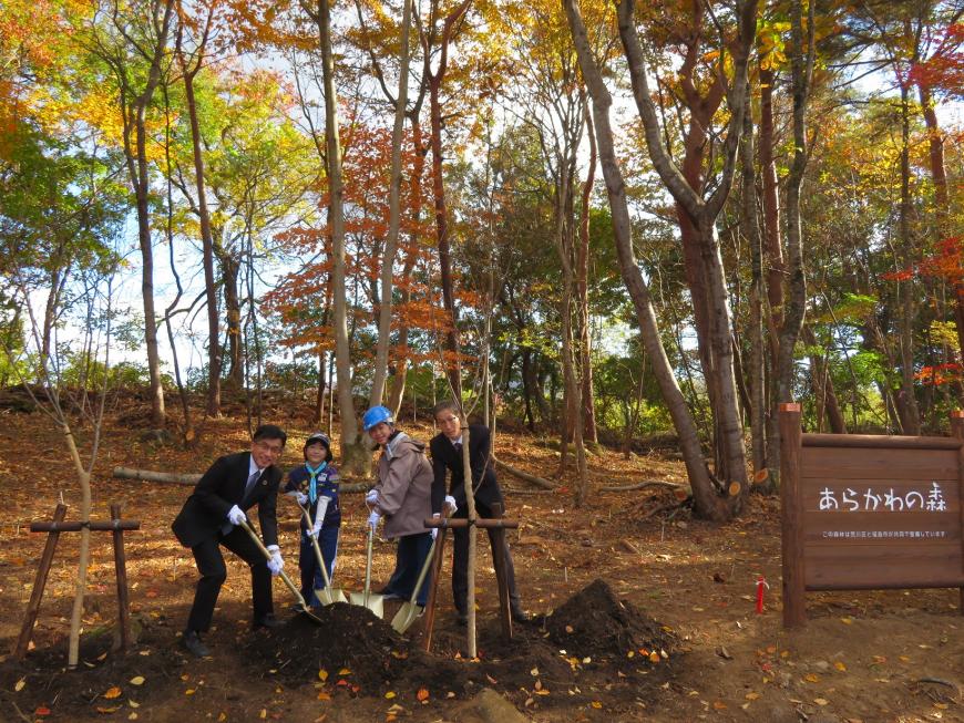 「あらかの森」と書かれた看板がある森で、市長とヘルメットを被った1名、スーツ姿の男性、ボーイスカウトの1名がそれぞれシャベルを持ち同じ場所を掘っている写真