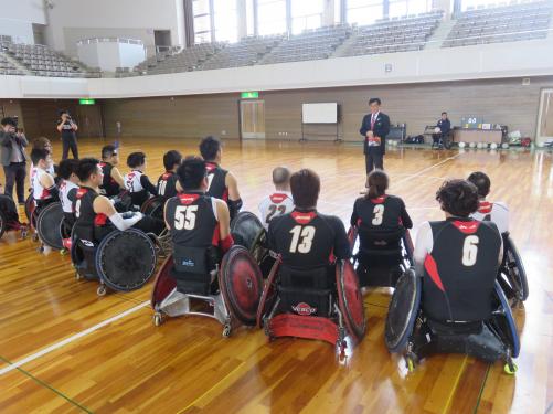 体育館の中で、ユニフォームを着て競技用の車いすに乗った選手たちが前に立った市長の方を向いている写真