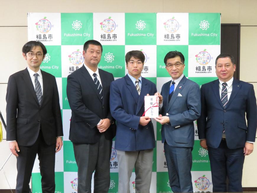 バックパネルの前に、生徒と関係者3名が立ち、市長が生徒に目録を手渡している写真