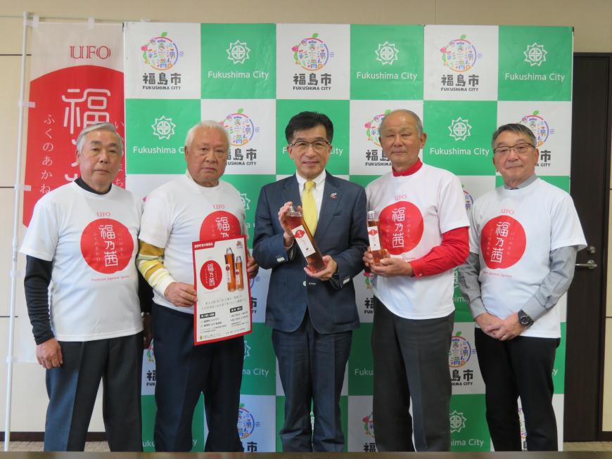 バックパネルの前に、市長の両隣に「福乃茜」の文字入りのTシャツを着た男性が2名ずつ立ち、市長と男性1名が梅酒、男性1名がポスターを持っている写真