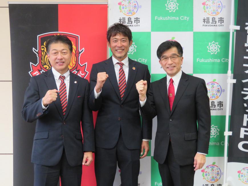バックパネルとチーム旗が並んでいる前で、市長と男性2名が右手でガッツポーズをしている写真