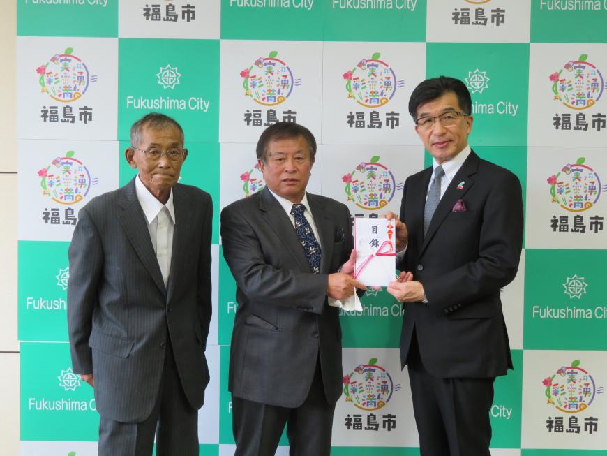 バックパネルの前で、市長と男性1名で目録を持ち、横に男性1名が立っている写真