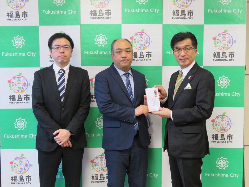 バックパネルの前に、市長を含む男性3名が立ち、市長と男性1人で目録を持っている写真