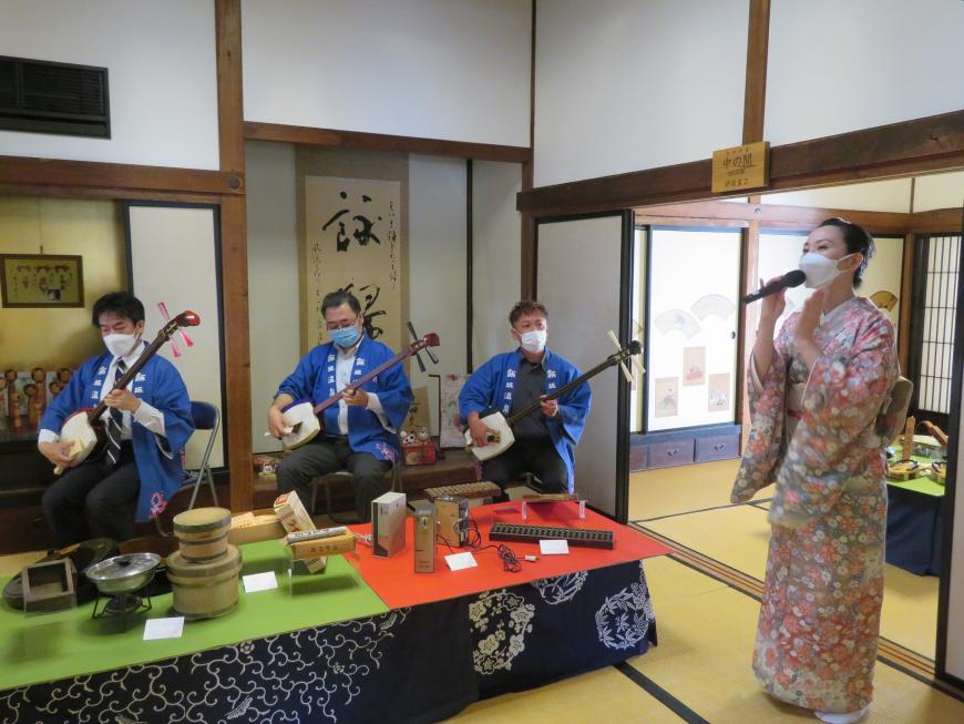 調度品が飾られた部屋で法被を着た3人の男性の三味線の演奏に合わせて着物姿の女性がマイクを持ち歌っている写真