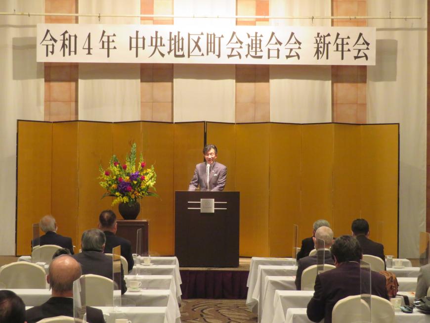 「中央地区町会連合会新年会」と書かれたボードがある会場で、座っている参加者の前で、市長が金屛風の前の演台に立って話をしている写真