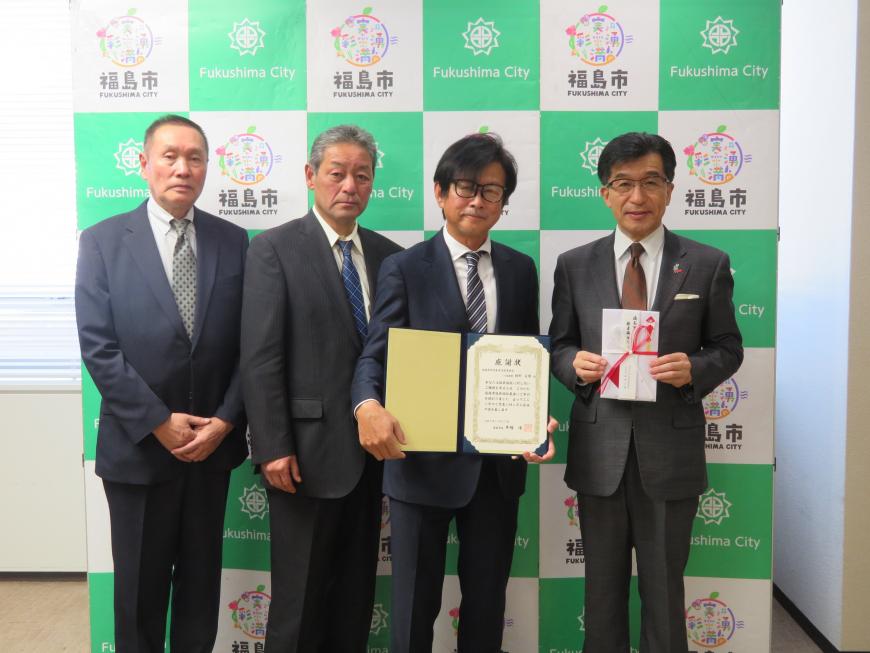 バックパネルの前に、市長を含む男性4名が立ち、市長が目録を持ち、男性1名が証書ファイルをひろげている写真