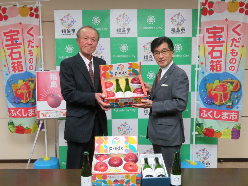 バックパネルの両脇に「くだものの宝石箱」と書かれた幟が立っている前で、市長と男性で4個のりんごと2本のシードルが入った箱を持っている写真