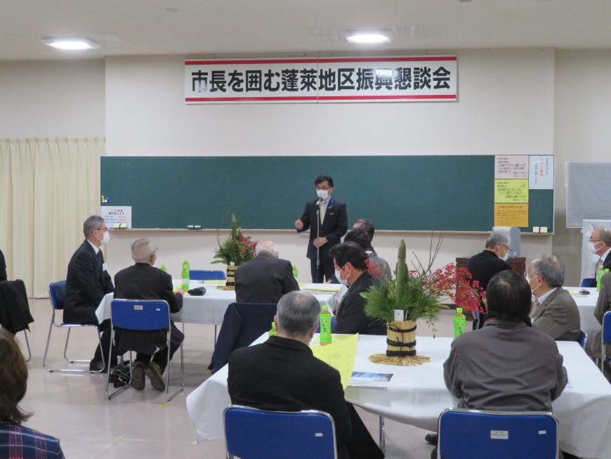 「市長を囲む蓬莱地区振興懇談会」と書かれたボードがある会場で、各テーブルに4人ぐらいに分かれて座っている参加者の前で、市長が立って話をしている写真