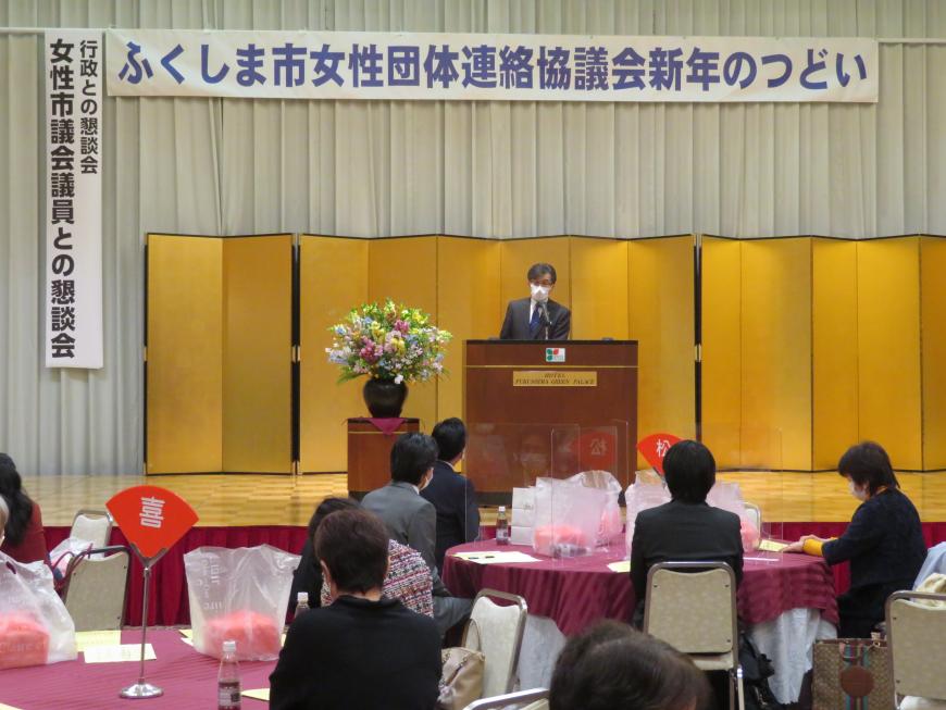 「ふくしま市女性団体連絡協議会新年のつどい」の横断幕がある会場で、円卓のテーブルに分かれて座っている参加者の前で、市長がステージの演台に立って話をしている写真