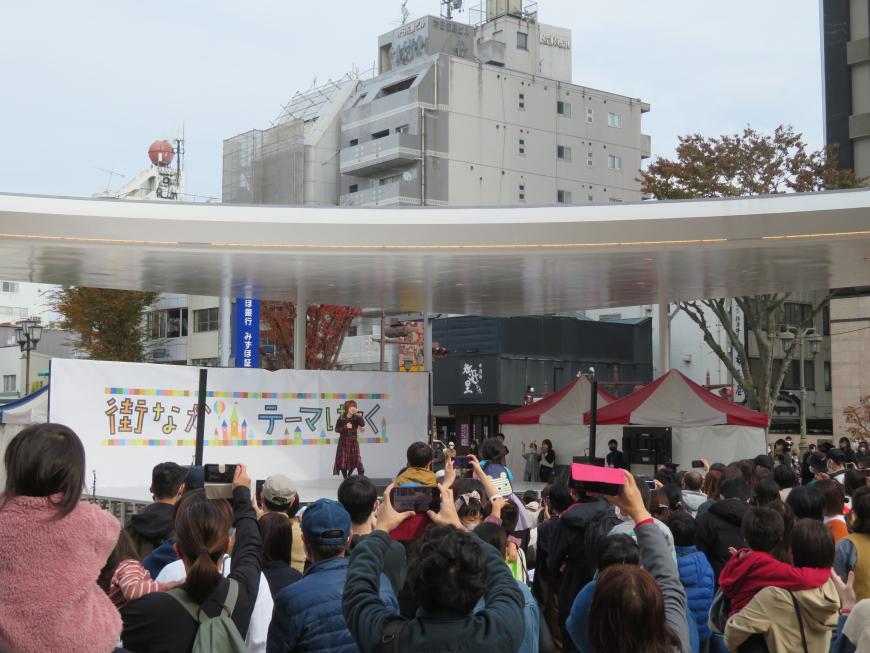 ステージに立つ女性を多くの観客の人がスマホで撮影しようとしているところを後方から撮影した写真