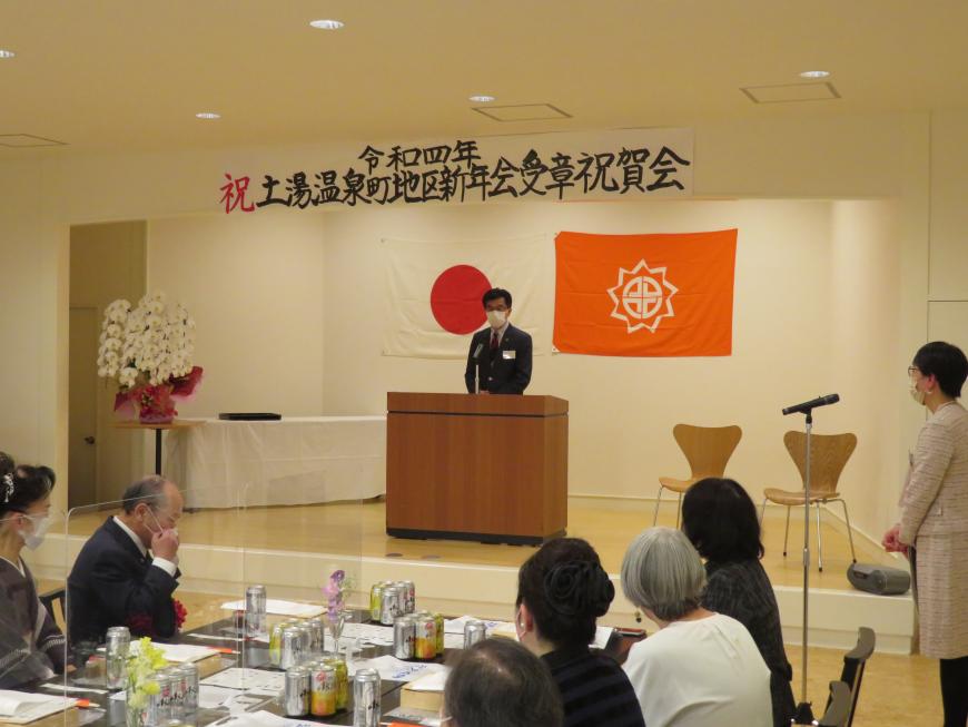 「土湯温泉町地区新年会受章祝賀会」の横断幕がある会場で、座っている参加者の前で、市長が演台に立って話をしている写真