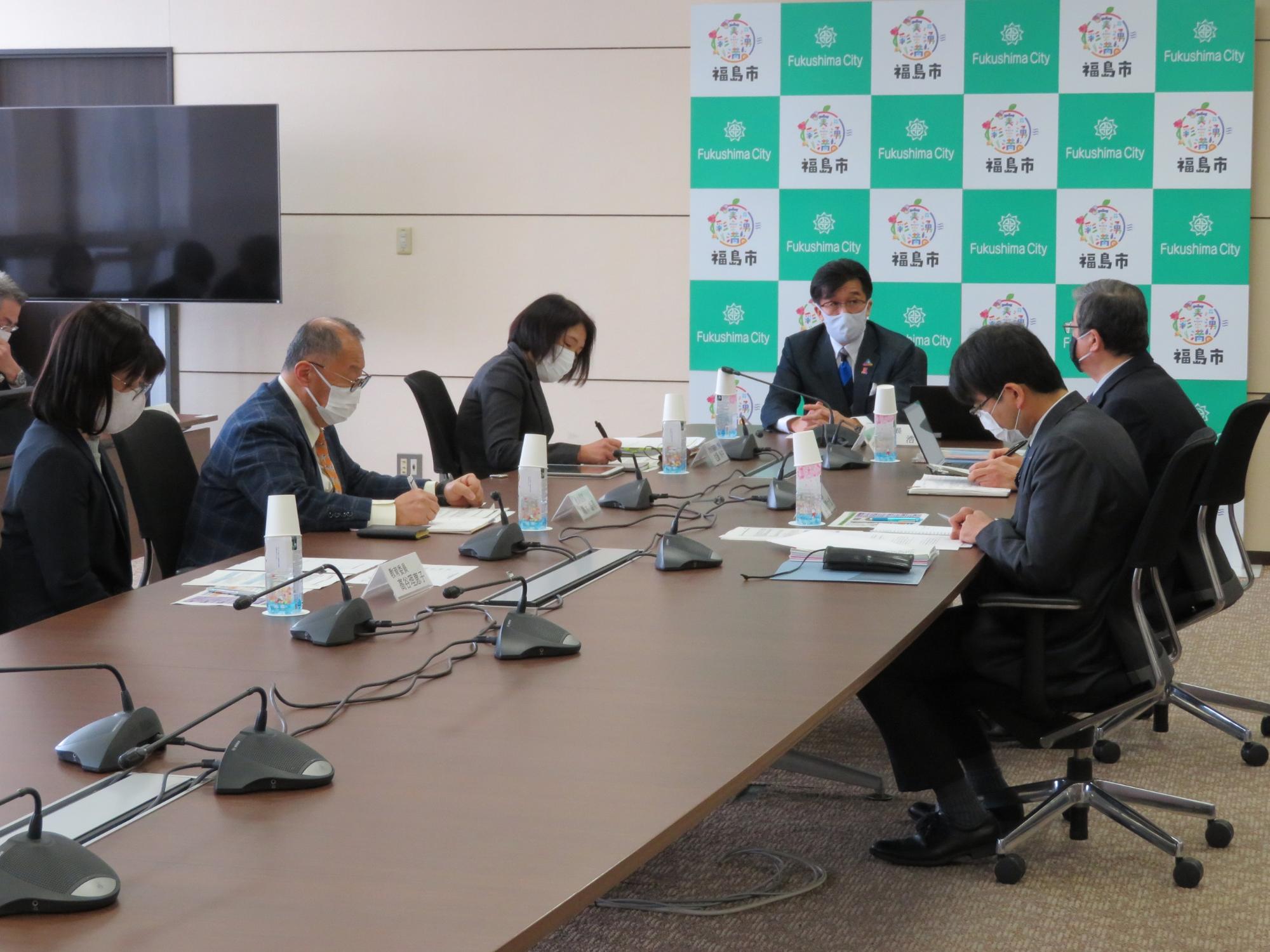 壁に福島市のバックパネルが設置された会議室で、市長と関係者が会議を行っている写真