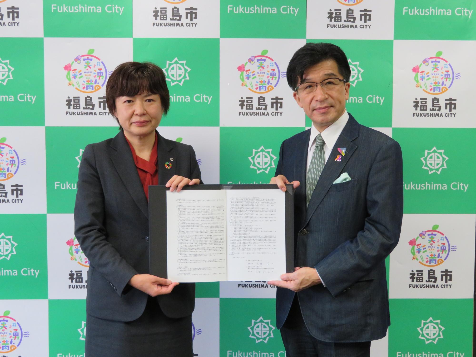 福島市のバックパネル前で、市長と東日本電信電話福島支店が協定書を手に持っている写真