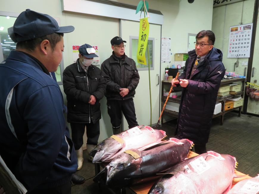 コートを着た市長が机に並べられている水揚げされた魚を見ながら業者の方と話をしている写真