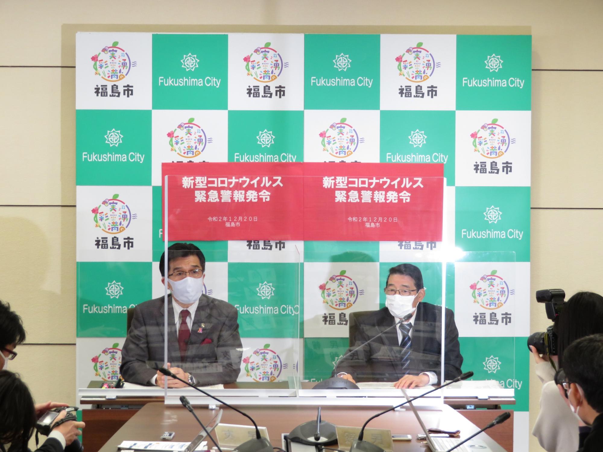 福島市のバックパネルの前に設けられた席に座った市長と関係者の男性が、「新型コロナ緊急警報発令」と書かれたパネルがついたアクリルボードを隔てて、集まった記者に会見を行っている写真