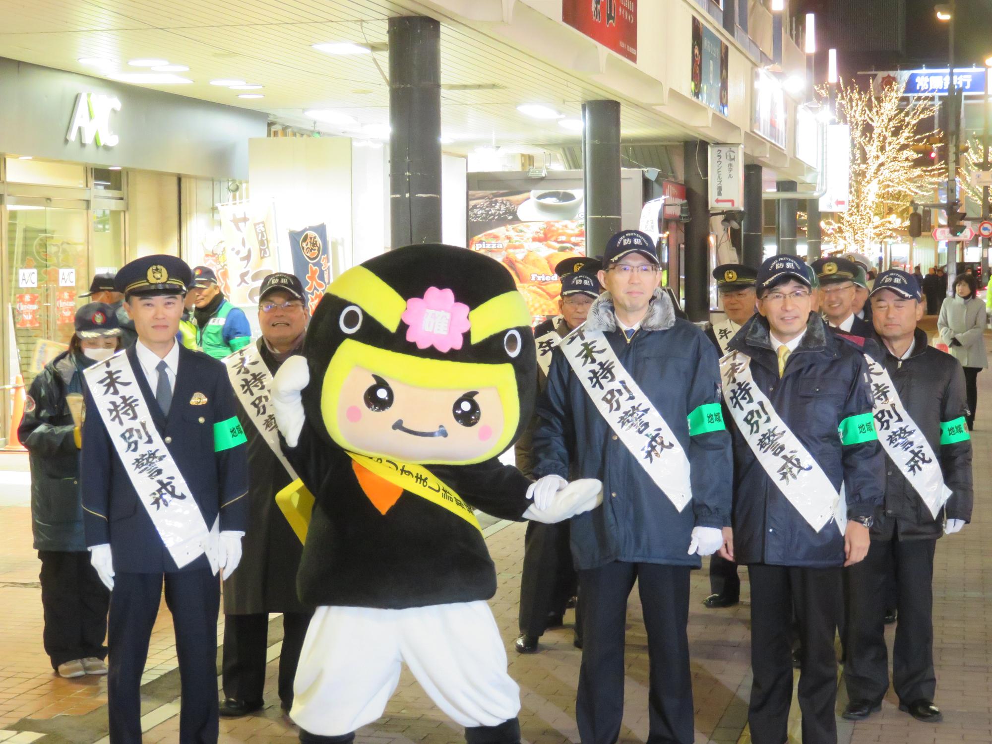 年末特別警戒の襷を掛けた警察官と市長、カクニンジャー福くんが駅前に集合している写真