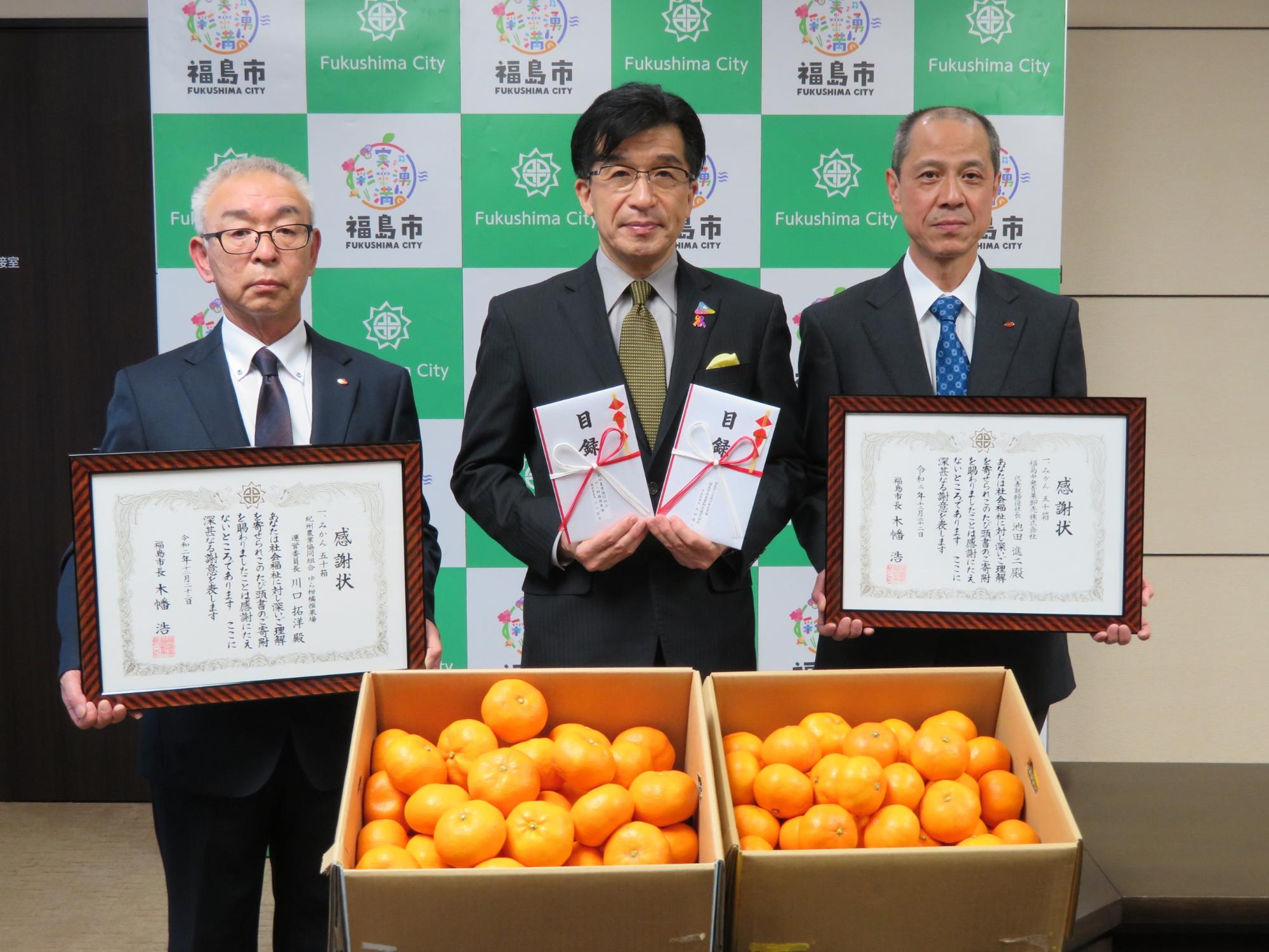 ふたつの箱いっぱいに詰まったみかんの前で、市長が2つの目録を、2名の関係者の男性が感謝状を手に持っている写真