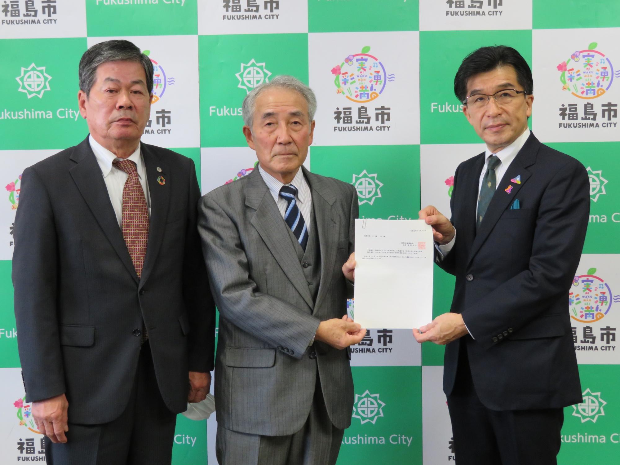 福島市のバックパネル前で、市長と二人並んで立った男性の一人が飯野地域審議会答申を両側から持っている写真