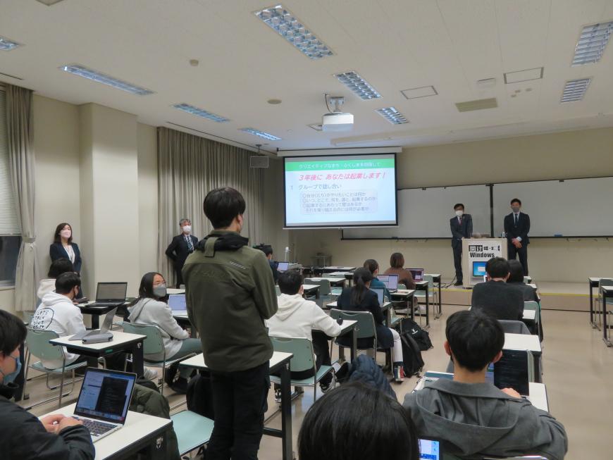 資料が映し出されたスクリーンが設置された室内で参加している学生が自席で立ち、前に立っている市長と男性に向かって話をしている写真