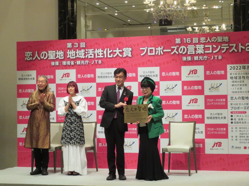 バックパネルの前で、市長と桂由美さんで「恋人の聖地」と書かれたプレートを持っている写真