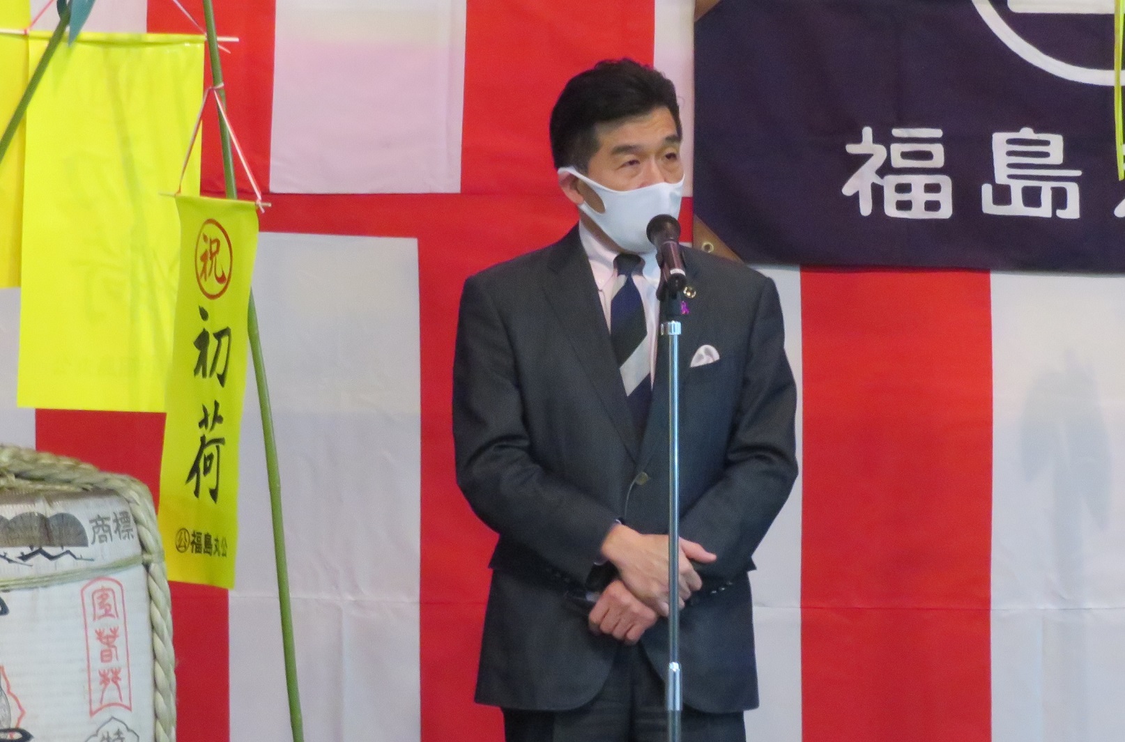市長がマイクスタンドの前で話をしており、背景には紅白幕が張られている写真