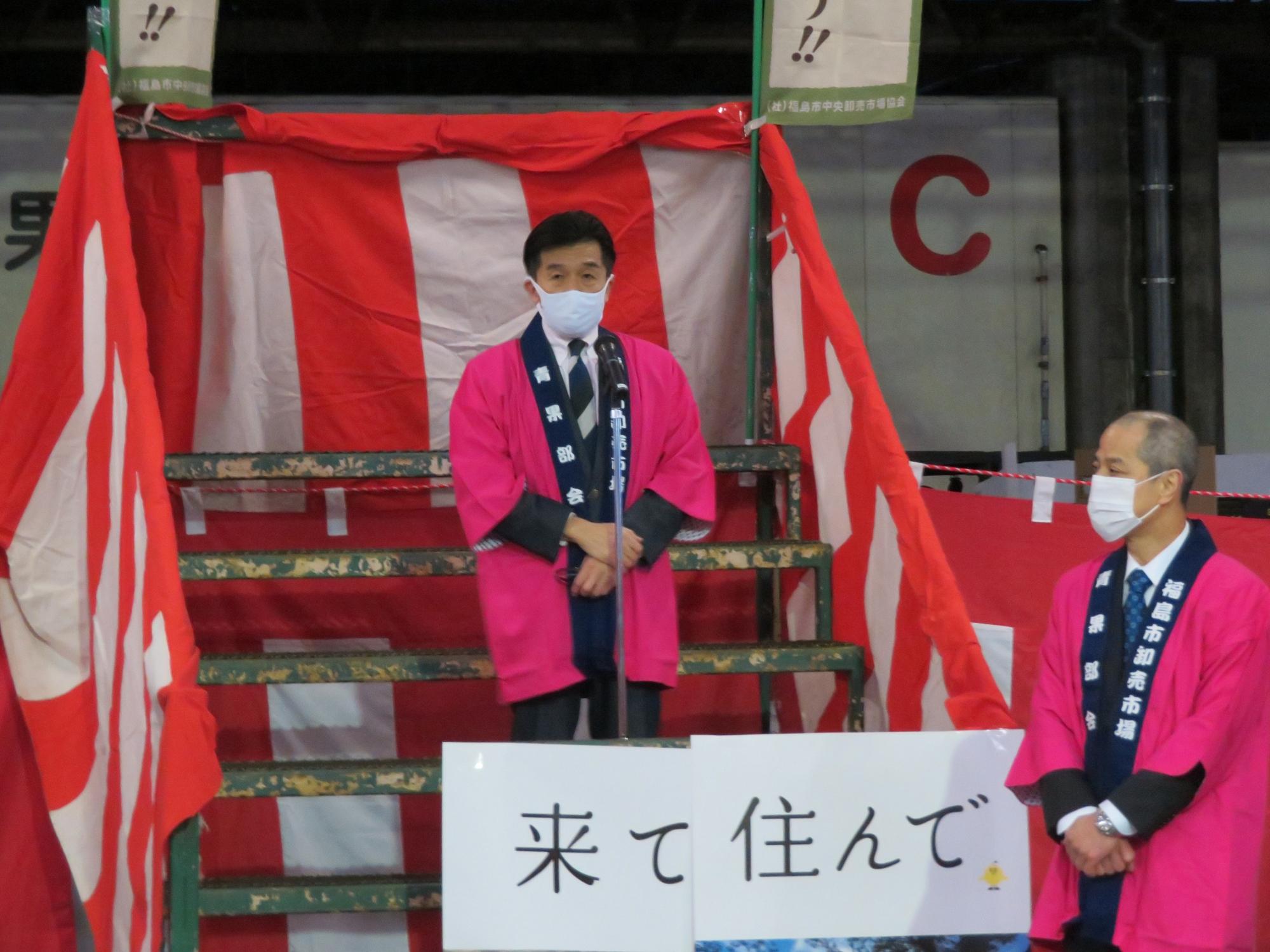 紅白幕の階段で、ピンク色のはっぴを着た市長がマイクスタンドの前で話をしている写真
