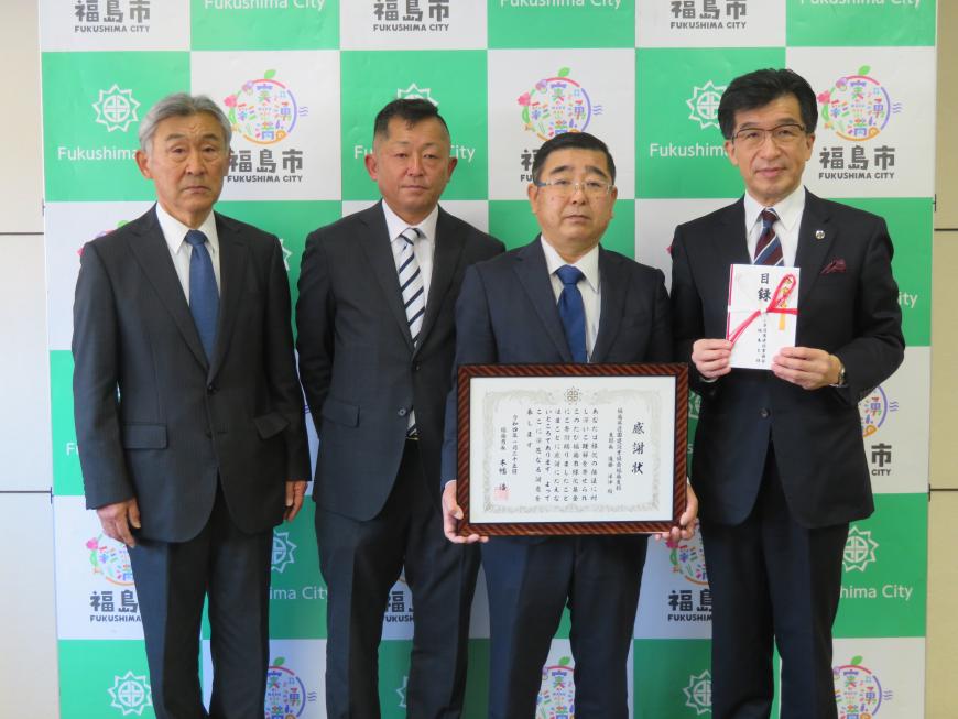 バックパネルの前で、市長が目録封筒、男性が額入りの感謝状をそれぞれ持ち、男性の横に2人の男性が立って並んでいる写真