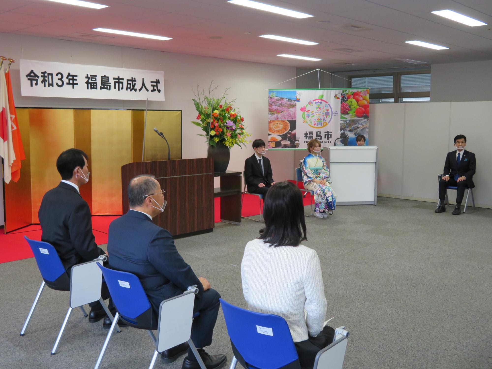 「令和3年度 福島市成人式」の吊りパネルがあり、金屏風の前にスーツ姿の男性と、振り袖姿の女性が座っており、市長と関係者が脇の方で椅子に座っている写真