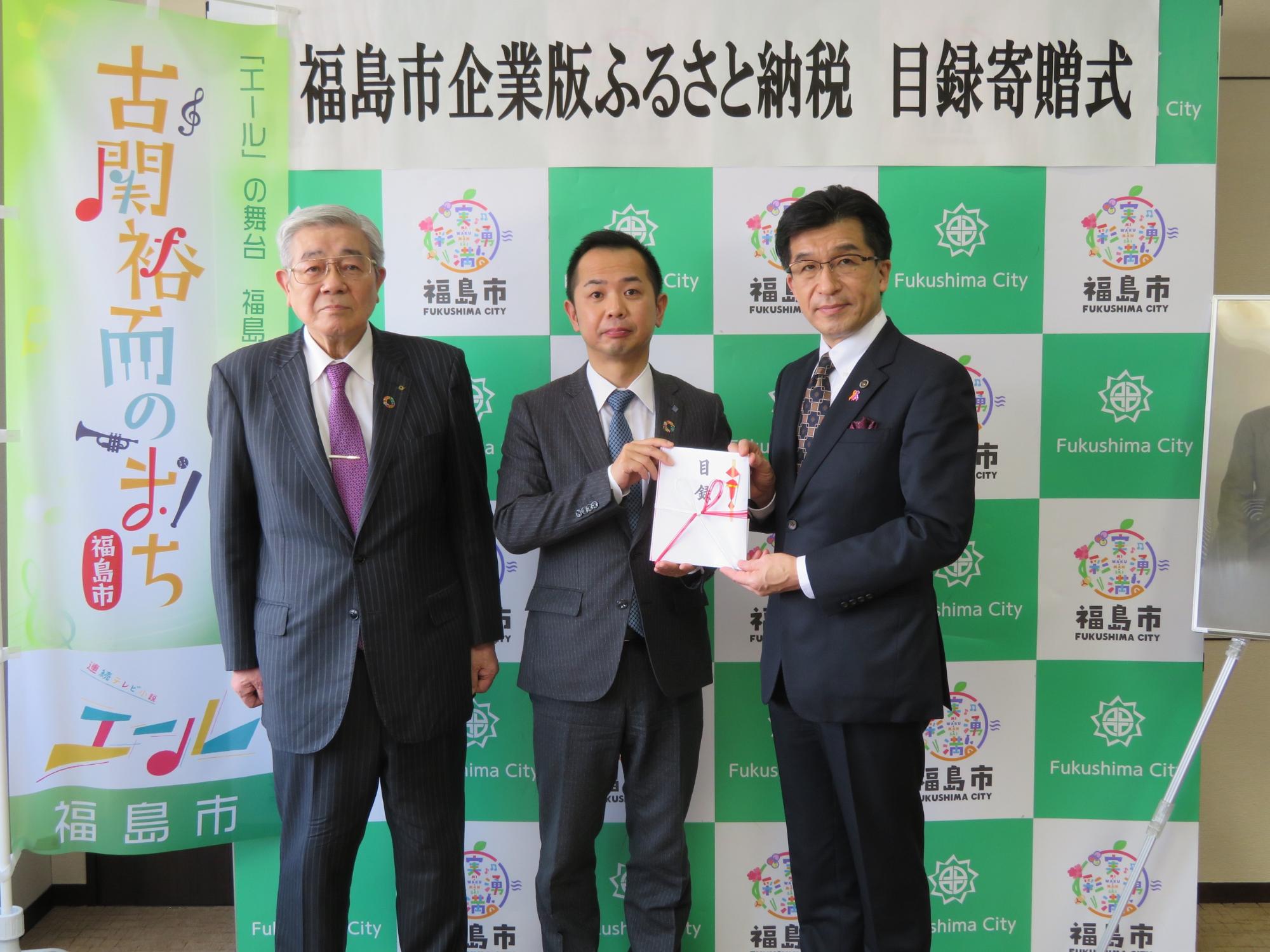 「福島市企業版ふるさと納税目録寄贈式」がパネルに貼られており、市長と関係者2名が並び、目録を二人で両手に持っている写真