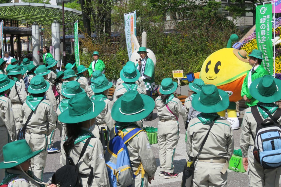 緑色のつば付きの帽子とグレーの制服を身に着けた子供たちがたくさん集まり、前方の緑色の帽子と法被を着た市長や黄色の丸いマスコットの方を見ている写真