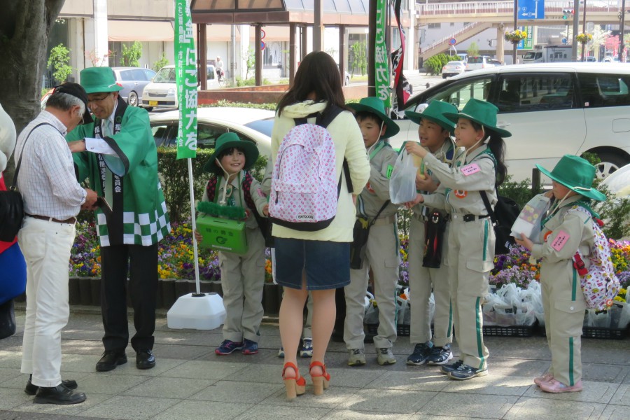 緑色のつば付きの帽子とグレーの制服を身に着けた市長や子供たちが、街頭に並んで通行人に募金を呼び掛けている様子の写真