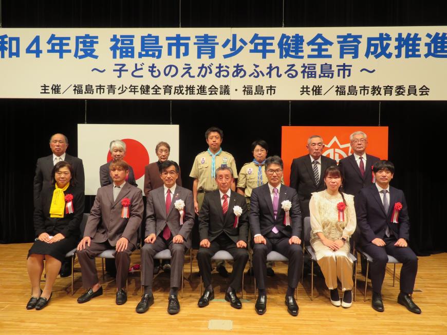 「令和4年度 福島市青少年健全育成推進大会」と書かれた横断幕と2枚の旗が掲げられたステージの前列に胸章リボンをつけた市長と表彰された6名が座り、後列に8名が立ち並んでいる記念写真
