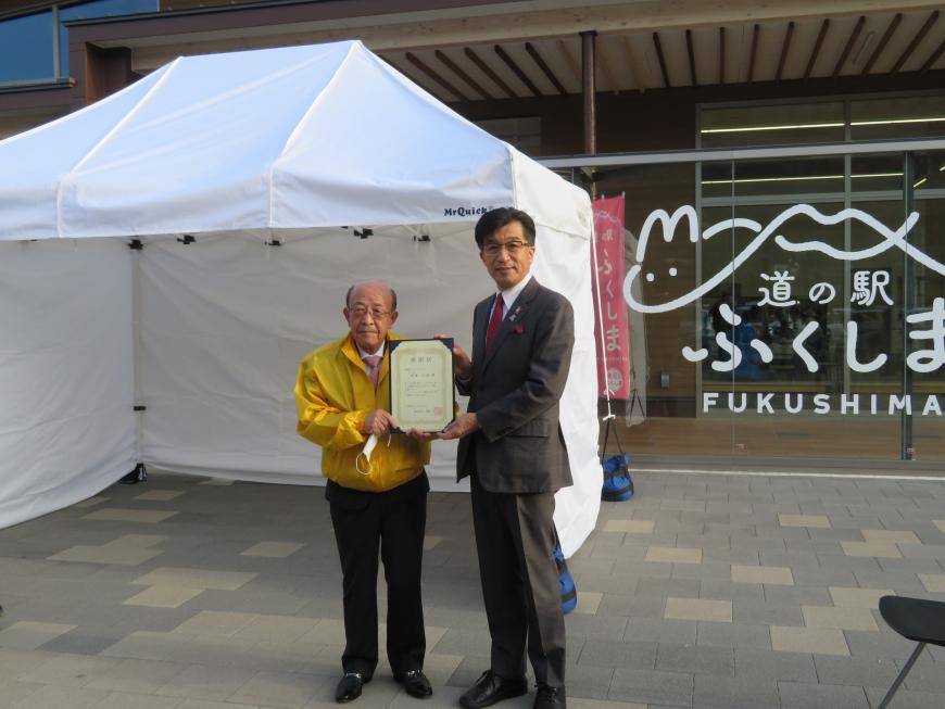 白いテントの前で市長と黄色の上着を着た男性で感謝状を持っている写真