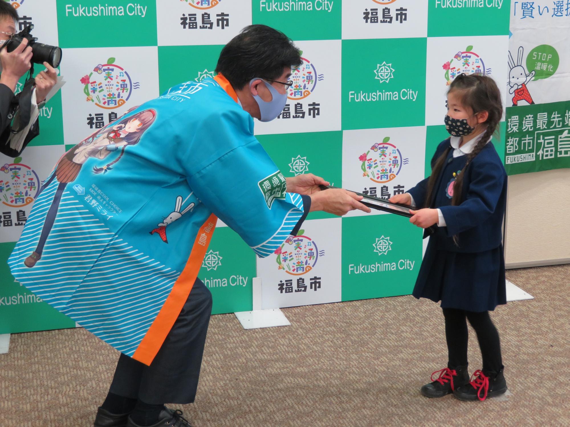 福島市のバックパネル前で、水色のはっぴを着た市長が、幼稚園生くらいの女の子と向き合い、中腰になって表彰状を手渡している写真