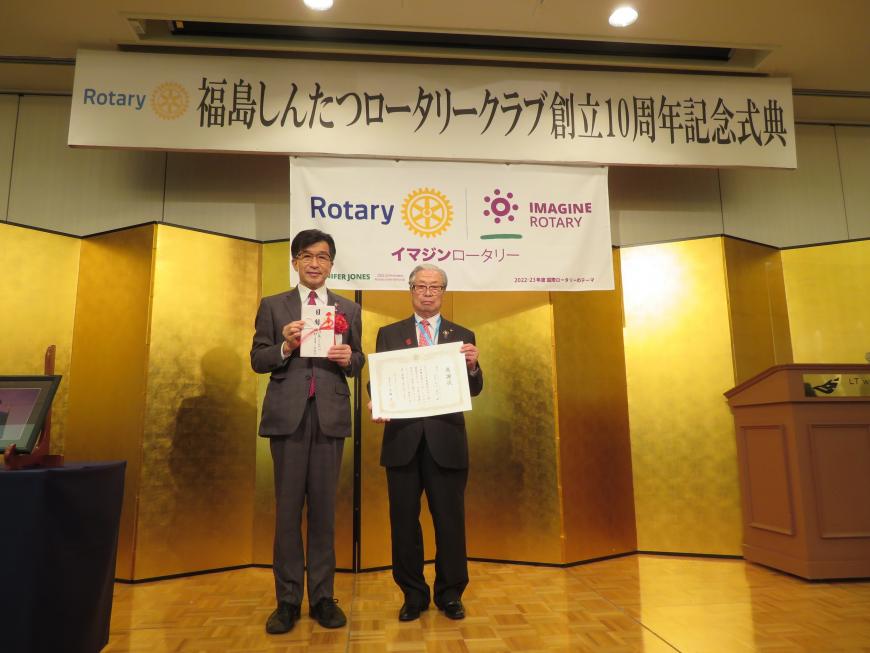 記念式典の金屛風の前で市長が目録を持ち、隣の男性が感謝状を持って並んでいる写真