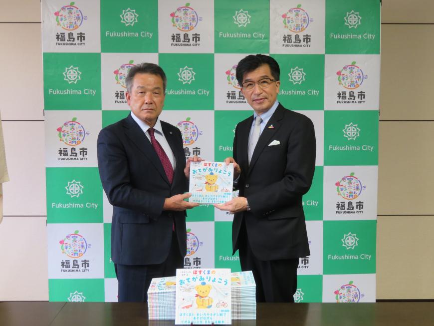 バックパネルの前で、市長と男性で絵本を持ちその前に寄付される絵本が机に置かれている写真