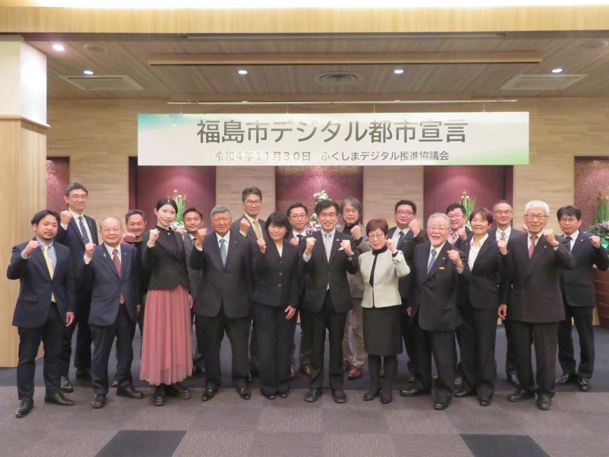 「福島市デジタル都市宣言」と書かれた横断幕をバックに、市長を含む20名ほどの参加者の方が横に2列に並んでガッツポーズをしている写真