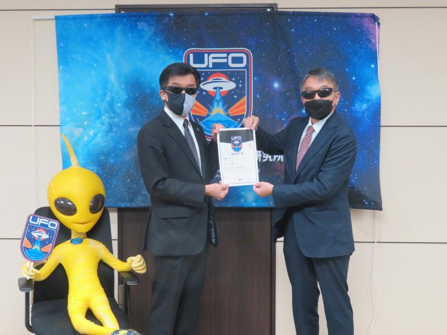 「UFO」の横断幕の前で、サングラスにマスク姿の市長と男性で任命書を持っている写真