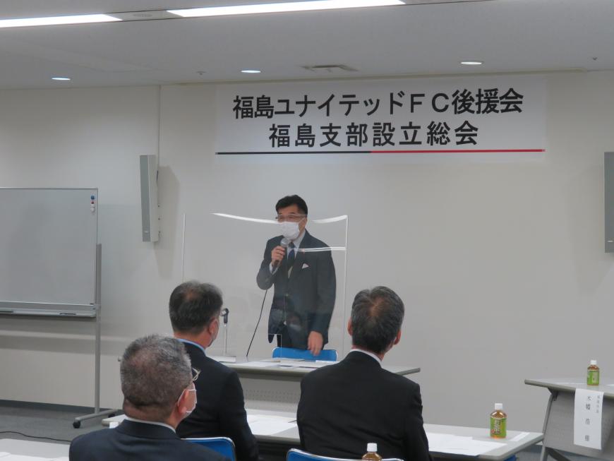 壁に「福島ユナイテッドFC後援会福島支部設立総会」と書かれた横断幕が貼られた部屋で、市長が座っている参加者の前で起立し話をしている写真