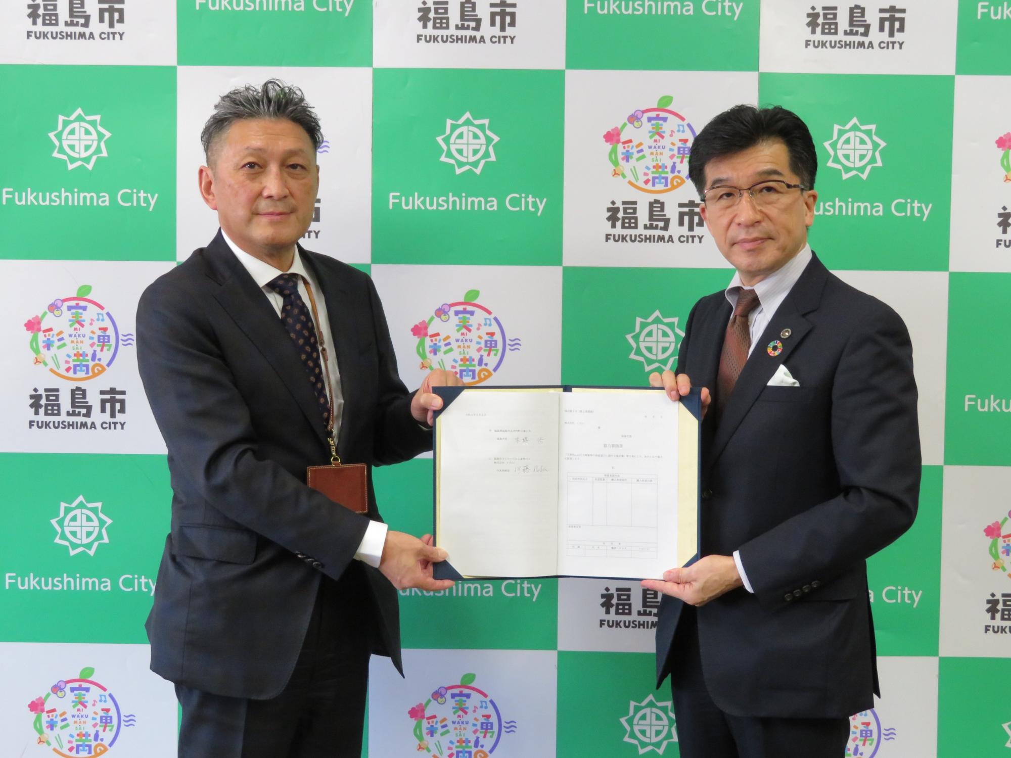 福島市のバックパネル前で、市長と株式会社いちいの関係者と一緒に協定書を持っている写真