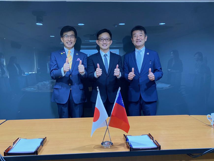 日本を台湾の国旗が置かれた机の後ろで市長を含む男性3名が両手で親指を立てたポーズをとっている写真