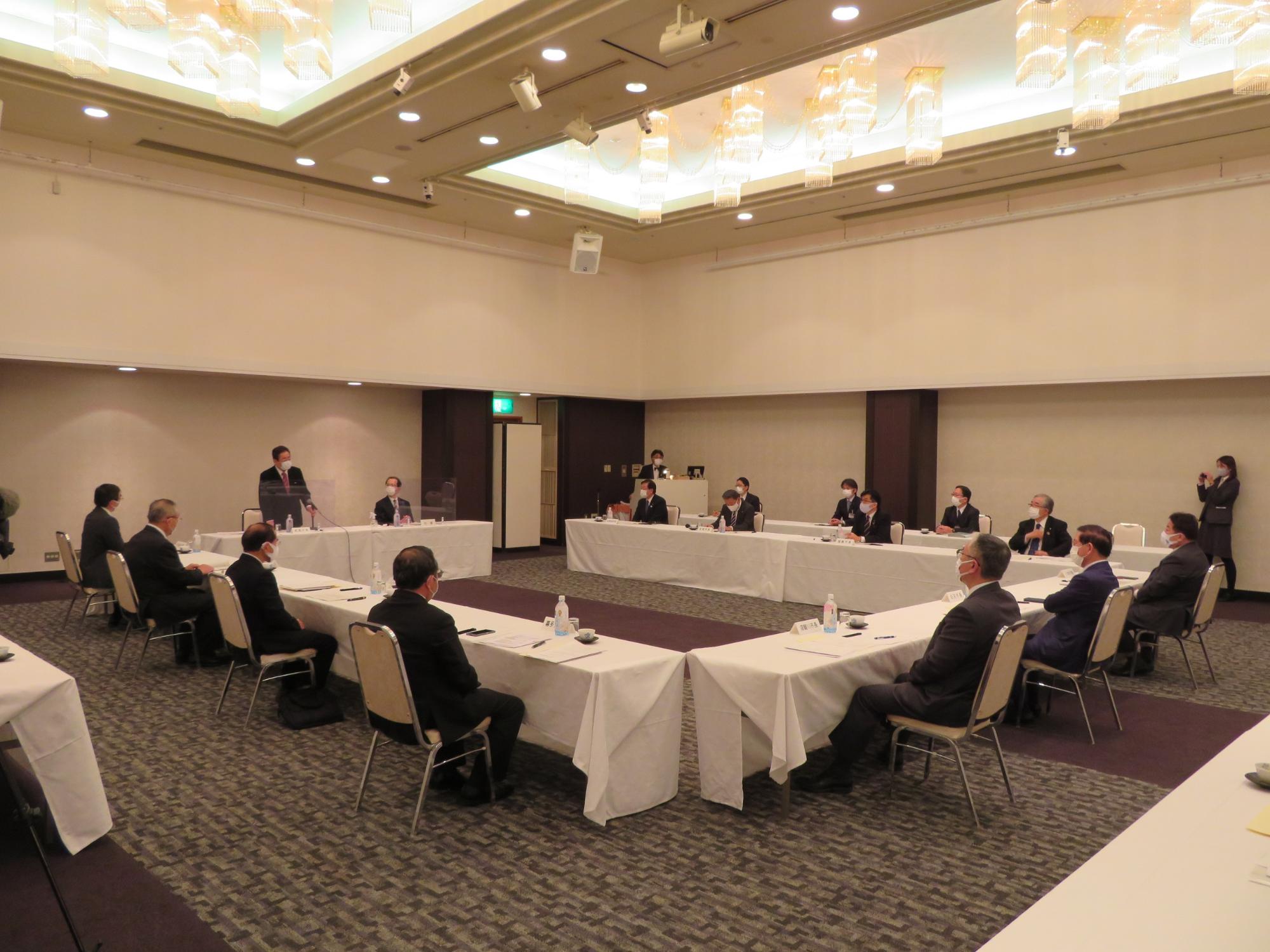 ホテルの広い会場で、コの字に並べられた机の席に関係者が椅子に座って意見交換を行っている写真