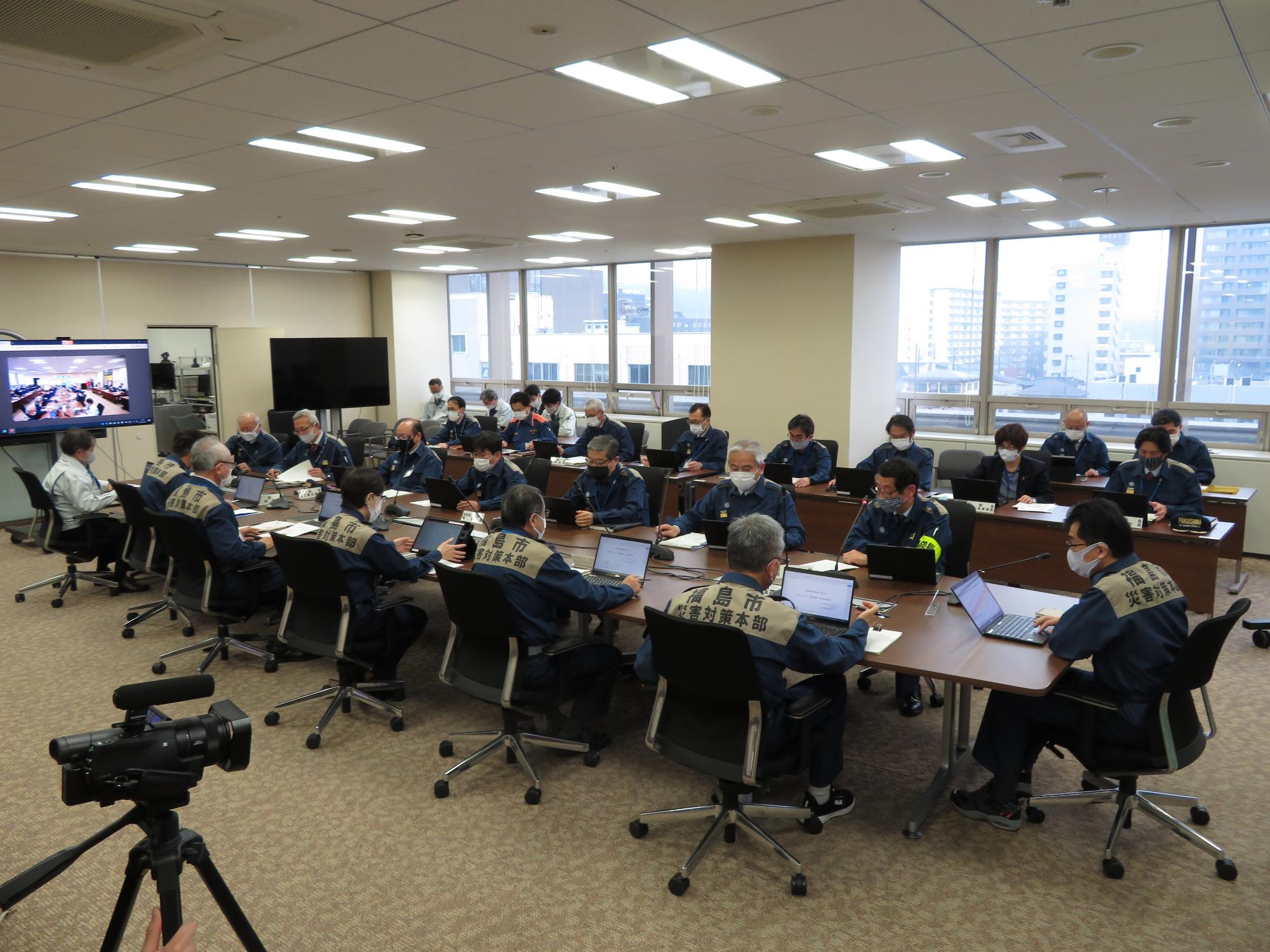 庁議室の一室で、市長と災害対策本部の関係者らが長机の席に座りパソコンを見ている写真