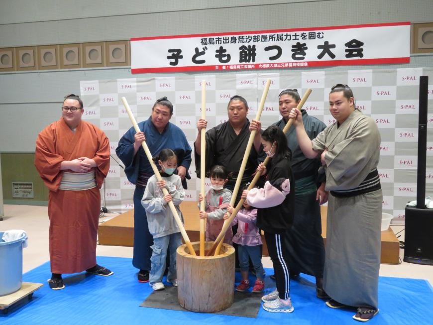 異なる色の着物を着た5名の力士のうち4名が、子どもたちが持ったそれぞれの棒の先を持ち、臼で餅をついている写真