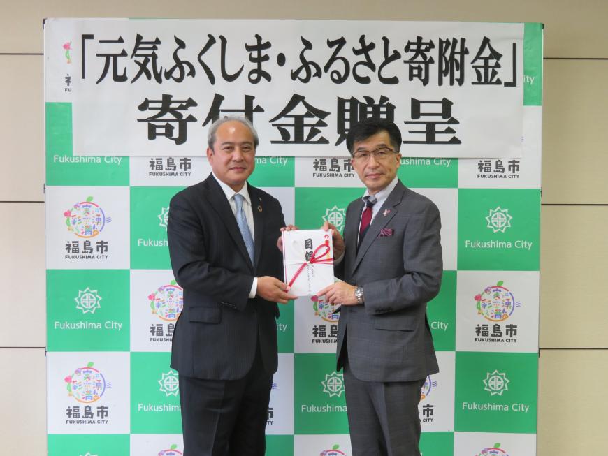 「元気ふくしま・ふるさと寄附金 寄付金贈呈」と書かれた紙が貼られたバックパネルの前で市長と対馬営業本部長が一緒に目録を持って記念撮影をしている写真