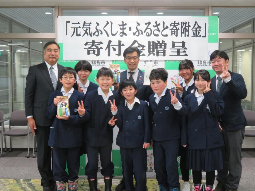 「寄附金贈呈」の横断幕の前で男性2名と児童7名が市長を囲むように立ち、ピースサインをしている写真
