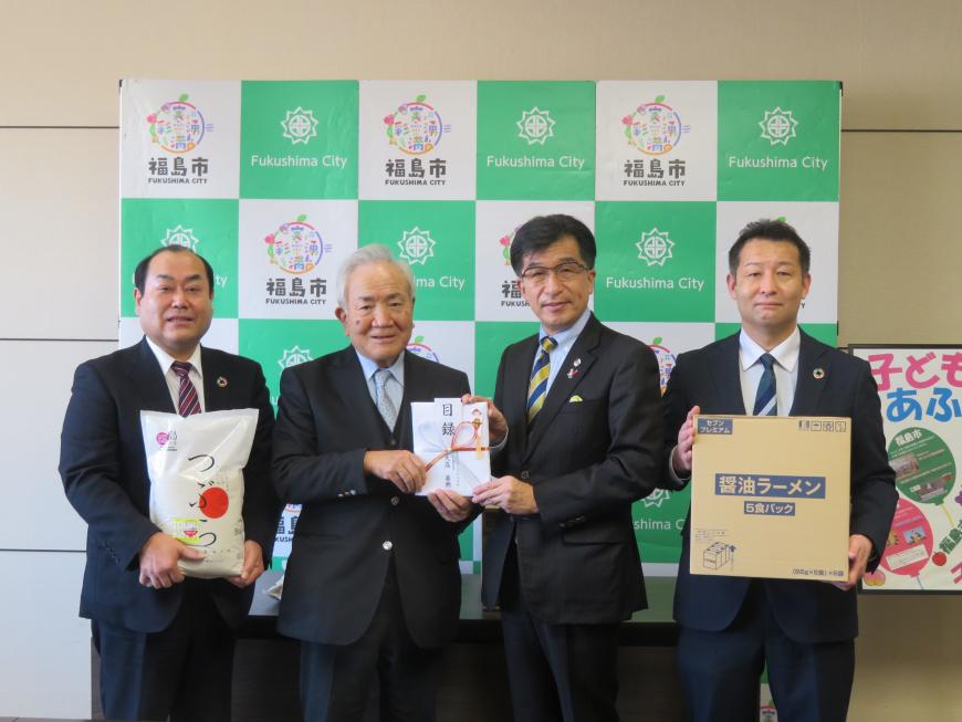 バックパネルの前で、市長と男性で目録を持ち、その両脇の男性がビニール袋に入ったお米と醤油ラーメンの箱をそれぞれ持っている写真