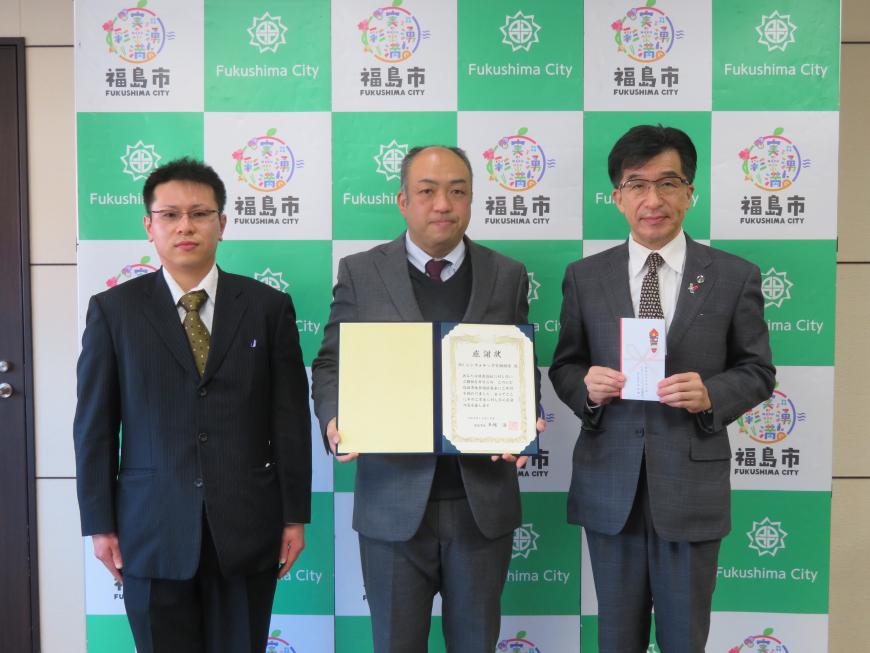 バックパネルの前で、市長の横に男性2名が立ち、1名が感謝状を持ち、市長が目録を持っている写真